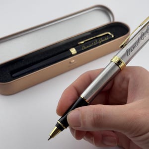 May include: A silver and black pen with gold accents, engraved with "Daniel C. Groth." The pen is held in a hand, with a matching pen in a gold-colored case. The case is open, revealing the pen inside.