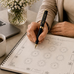 May include: A black and gold pen with the name "Albert Cutler" engraved on it is held over a notebook with a wedding reception seating chart. A cup of coffee, a laptop, and a vase of white flowers are also on the table.