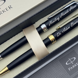 May include: Two black Parker pens with silver and gold accents, presented in a gift box. One pen reads "Healing Hands Hero" and the other reads "Dr. Adam Brantley". The pens are secured with a beige ribbon.