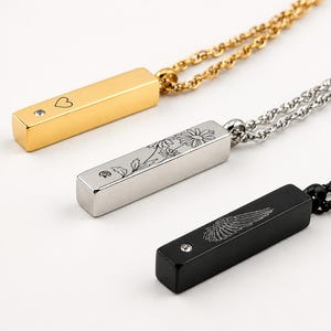 May include: Three bar pendant necklaces in gold, silver, and black tones. Each rectangular pendant features a small embedded crystal and a unique engraving: a heart, a daisy floral design, and a single wing, all suspended on matching chains.