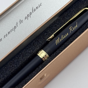 May include: A black pen with gold accents and the name "Melissa Reed" engraved in gold lettering. The pen is presented in a gold-colored box with the phrase "From concept to applause" printed inside.