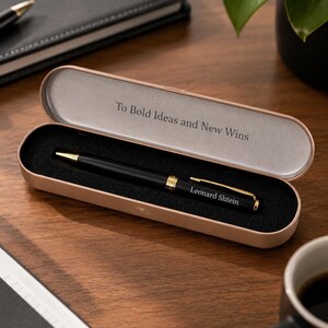 May include: A black ballpoint pen with gold-colored accents and the name Leonard Shtein engraved on the barrel, resting inside a rose gold-colored metal case with a black foam insert. The interior lid features the text To Bold Ideas and New Wins.