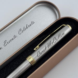May include: A silver pen with gold accents and the name "Carol McCollum" engraved on the side, presented in an open, bronze-colored metal case. The case lid reads "Plan. Execute. Celebrate."