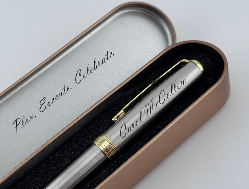 May include: A silver pen with gold accents and the name "Carol McCollum" engraved on the side, presented in an open, bronze-colored metal case. The case lid reads "Plan. Execute. Celebrate."
