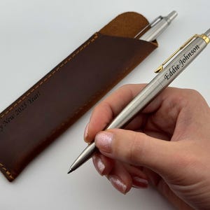 May include: A silver pen with a gold accent and the name "Eddie Johnson" engraved on it. The pen is in a brown leather case with the text "Happy New 2023 Year!" engraved on it.