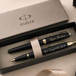May include: A Parker brand gift box containing two matte black pens with gold-tone accents. The top pen is engraved with the name Ryan Boileau, and the bottom pen is engraved with the text You Taught Me Well. Both pens are held by a beige ribbon.