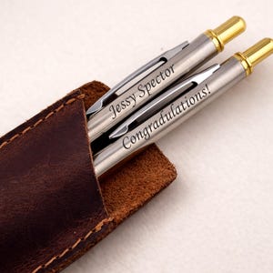 May include: Two silver pens with gold accents, engraved with "Jessy Spector" and "Congratulations!" are displayed in a brown leather pen case. The pens are a sleek, professional design, perfect for gifting or personal use.