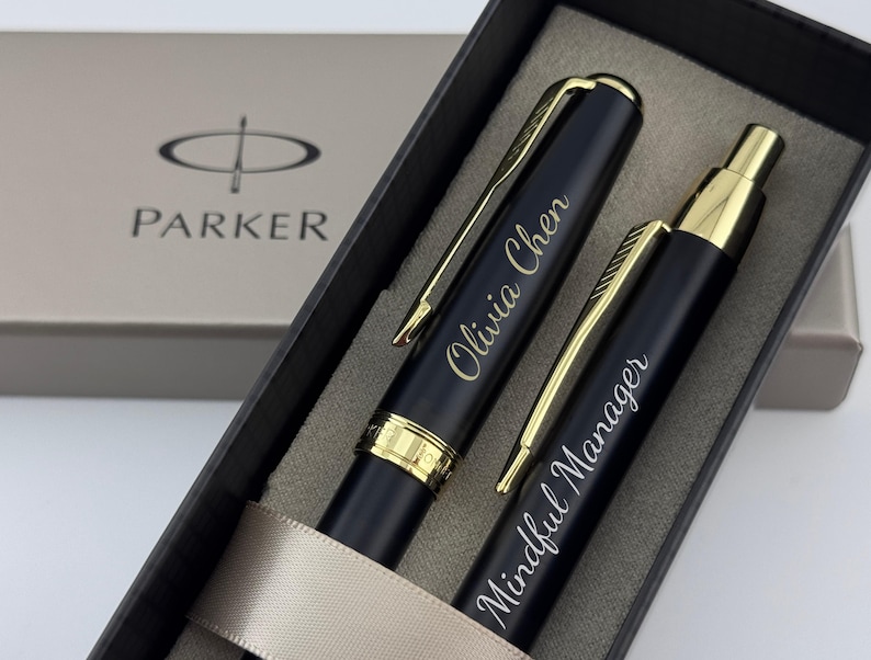 May include: A Parker pen set in a black presentation box. The set includes a fountain pen and a ballpoint pen, both in matte black with gold-tone accents. The fountain pen is engraved with Olivia Chen, and the ballpoint pen with Mindful Manager.