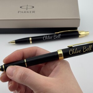 May include: A black ballpoint pen with gold-colored accents and the name Chloe Bell engraved in gold script. The pen features a gold clip and a central gold band. A matching pen and a Parker branded gift box are visible in the background.