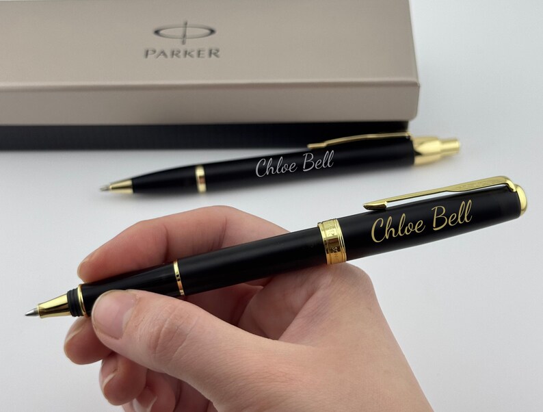 May include: A black ballpoint pen with gold-colored accents and the name Chloe Bell engraved in gold script. The pen features a gold clip and a central gold band. A matching pen and a Parker branded gift box are visible in the background.