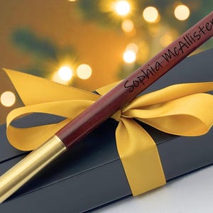 May include: A personalized pen with the name "Sophia McAllister" engraved on the dark wood barrel. The pen has a gold-colored tip and end cap. It is presented in a dark gray gift box and tied with a yellow ribbon.
