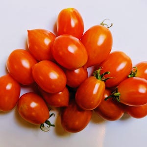 May include: A pile of ripe, oval-shaped tomatoes with vibrant red-orange skin and green stems. The tomatoes are clustered together, showcasing their smooth, glossy texture. The image is taken from a high angle, highlighting the freshness of the produce.