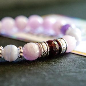 Puede incluir: Una pulsera de cuentas que presenta una secuencia de piedras en tonos violeta claro, lavanda y azul pálido. El diseño incluye una cuenta facetada de color rojo oscuro y varios espaciadores de metal plateado con patrones florales.