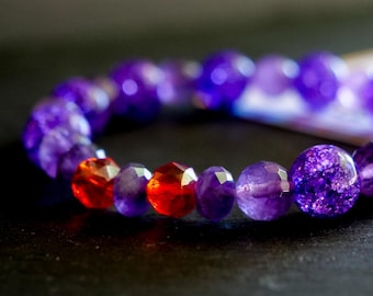 Puede incluir: Una pulsera de cuentas con una mezcla de cuentas redondas de amatista morada, cuentas de vidrio morado facetadas y dos cuentas facetadas de color naranja vibrante. La pulsera incluye una cuenta de vidrio morado craquelado y texturizado.
