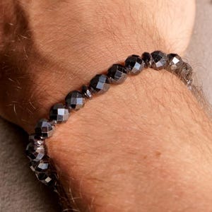 Puede incluir: Una pulsera y un collar compuestos por pequeñas cuentas oscuras, facetadas y de aspecto metálico. La pulsera se lleva en la muñeca, mientras que el collar cuelga, ambos con una superficie pulida y reflectante de forma circular uniforme.