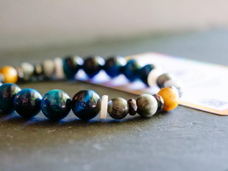 Puede incluir: Una pulsera de cuentas con esferas pulidas en tonos azul profundo, amarillo dorado y gris apagado. El dise&ntilde;o incorpora peque&ntilde;os espaciadores circulares blancos y planos, junto con cuentas met&aacute;licas oscuras y facetadas intercaladas.