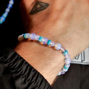 May include: A beaded bracelet with a mix of light blue, turquoise, and iridescent beads. The bracelet is worn on a wrist, with a black jacket sleeve visible. The beads vary in size and shape, creating a unique accessory.