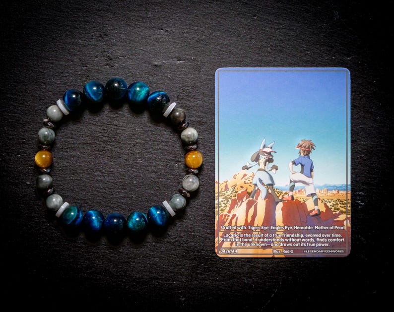 Puede incluir: Una pulsera de cuentas con piedras de ojo de tigre azul, hematita gris y ojo de tigre amarillo con espaciadores blancos, junto a una tarjeta coleccionable rectangular que muestra un personaje y una persona en un paisaje des&eacute;rtico rocoso.