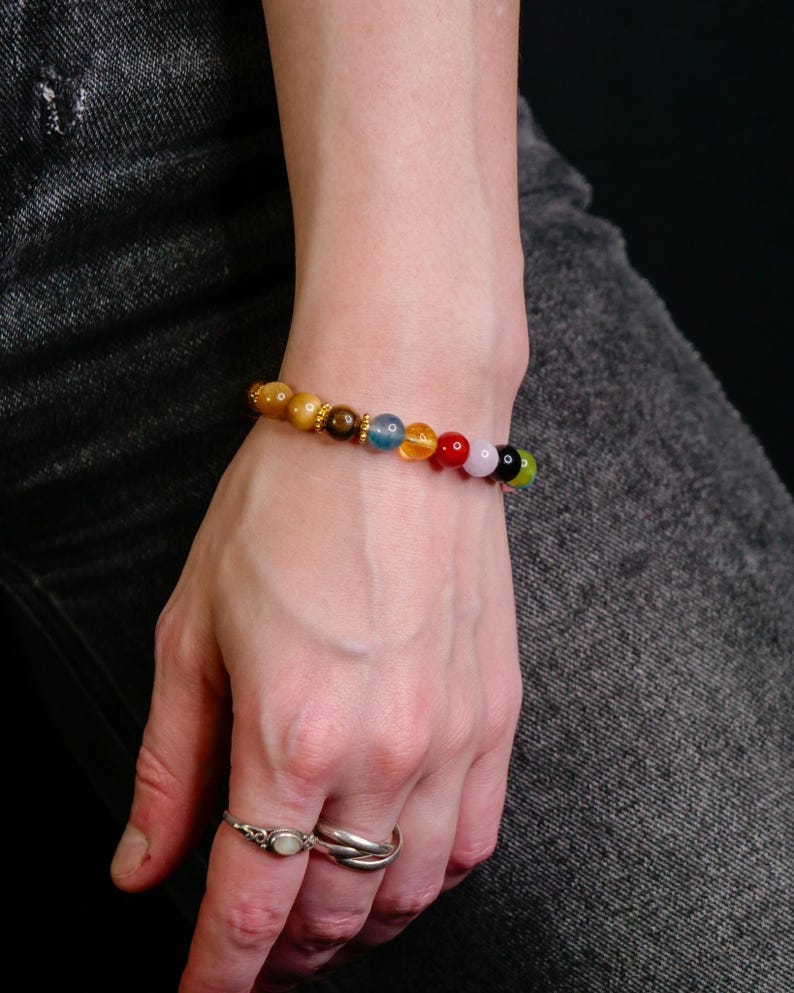 May include: A beaded bracelet featuring a sequence of round, polished stones in shades of yellow, brown, blue, orange, red, pink, black, and green, accented with small gold-toned metal spacers, worn on a wrist alongside two silver-toned rings.