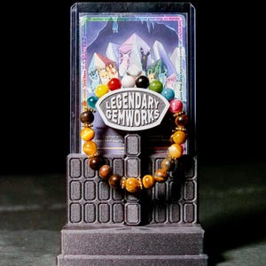 Puede incluir: Una pulsera de cuentas con una mezcla de piedras preciosas amarillas, marrones y multicolores con espaciadores dorados. La pulsera se exhibe en un soporte gris oscuro con un letrero que dice LEGENDARY GEMWORKS, frente a una tarjeta.