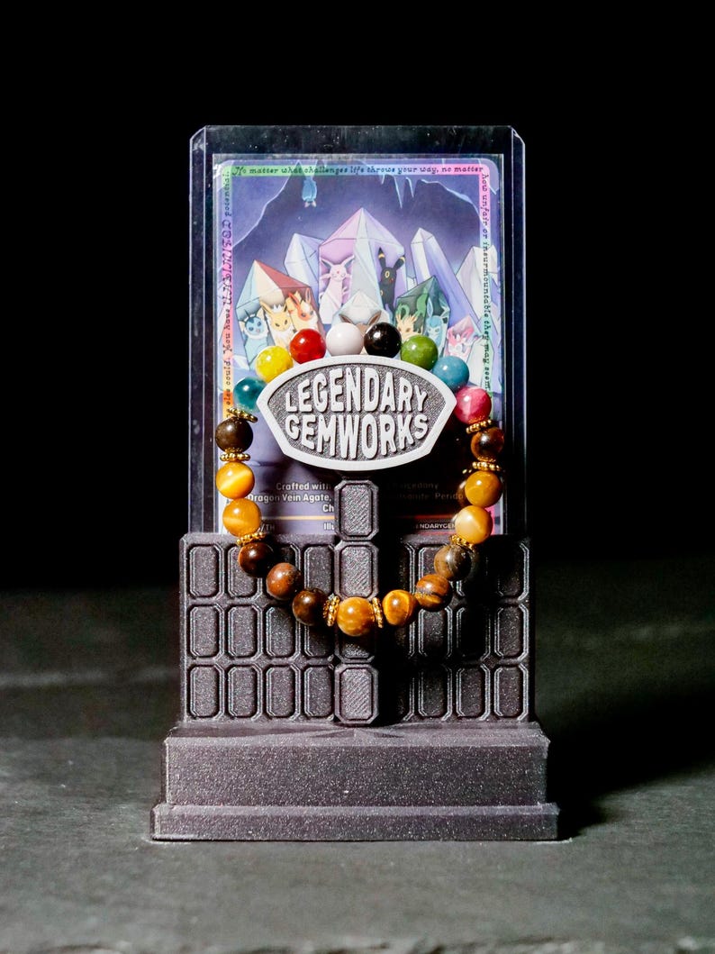 Puede incluir: Una pulsera de cuentas con una mezcla de piedras preciosas amarillas, marrones y multicolores con espaciadores dorados. La pulsera se exhibe en un soporte gris oscuro con un letrero que dice LEGENDARY GEMWORKS, frente a una tarjeta.