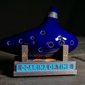 May include: A dark blue, glossy ocarina with several finger holes, displayed on a grey stand. The stand has the text "OCARINA OF TIME" in blue. The ocarina has a triangular emblem on its top and the stand.