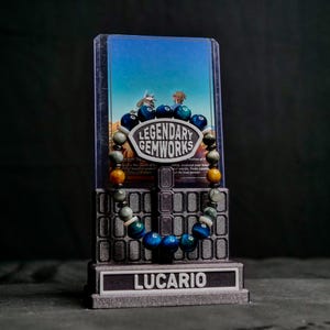 Puede incluir: Una pulsera de cuentas con piedras preciosas azules, verdes y amarillas sobre un soporte. El soporte tiene una placa central que dice LEGENDARY GEMWORKS y una base con la etiqueta LUCARIO. Detr&aacute;s de la pulsera hay una tarjeta con un cielo azul.
