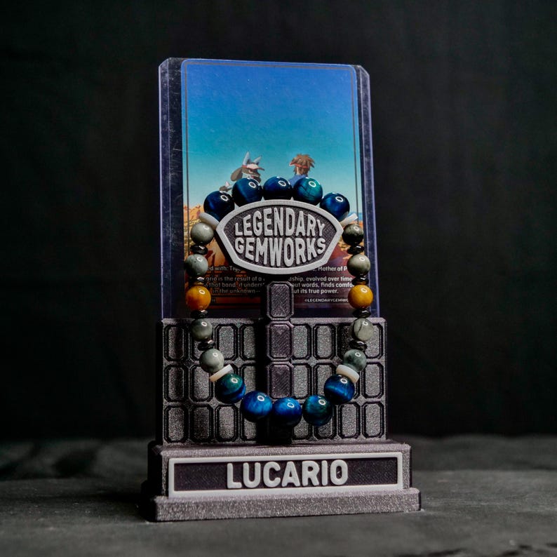 Puede incluir: Una pulsera de cuentas con piedras preciosas azules, verdes y amarillas sobre un soporte. El soporte tiene una placa central que dice LEGENDARY GEMWORKS y una base con la etiqueta LUCARIO. Detr&aacute;s de la pulsera hay una tarjeta con un cielo azul.