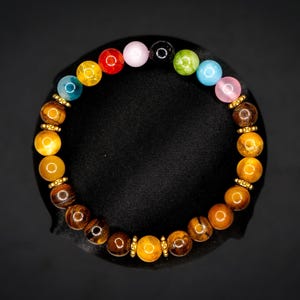 Puede incluir: Una pulsera de cuentas con una disposici&oacute;n circular de piedras ojo de tigre pulidas en tonos marr&oacute;n y dorado, intercaladas con peque&ntilde;os espaciadores de metal dorado y una secci&oacute;n superior de cuentas redondas lisas multicolores en rojo, amarillo, azul, rosa, negro, verde y azul claro.