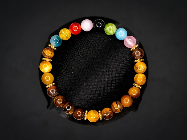 Puede incluir: Una pulsera de cuentas con una disposici&oacute;n circular de piedras ojo de tigre pulidas en tonos marr&oacute;n y dorado, intercaladas con peque&ntilde;os espaciadores de metal dorado y una secci&oacute;n superior de cuentas redondas lisas multicolores en rojo, amarillo, azul, rosa, negro, verde y azul claro.