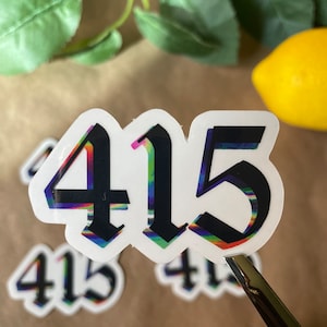 May include: A close-up of a sticker with the number 415 in a black gothic font. The numbers have a rainbow-colored glitch effect.