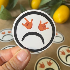 May include: A white sticker with a black outline depicting a cartoon face with a frown and two orange hands making the 'rock on' gesture.