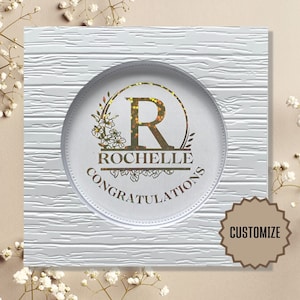 May include: White card with a woodgrain texture and a gold circle with a floral design and the letter "R" in the center. The text "ROCHELLE CONGRATULATIONS" is below the letter. The word "CUSTOMIZE" is on a brown starburst sticker in the lower right corner.