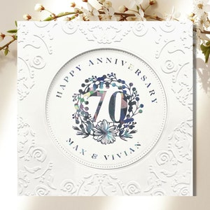 May include: White card with an embossed floral design. A circle in the center has the text "Happy Anniversary" and "Max & Vivian" with the number "70" in a colorful floral wreath.