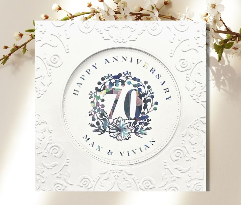May include: White card with an embossed floral design. A circle in the center has the text "Happy Anniversary" and "Max & Vivian" with the number "70" in a colorful floral wreath.
