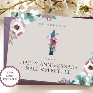 May include: A personalized anniversary card with floral accents. The card features the text "CELEBRATING 1 YEAR HAPPY ANNIVERSARY DALE & MICHELLE". The design includes watercolor flowers in shades of purple, teal, and pink. A sticker indicates two sizes are available.