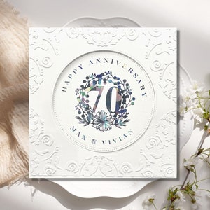 May include: White card with a decorative border and a circle in the center. The circle has a floral wreath with the number 70 in the center. The text "Happy Anniversary Max & Vivian" is below the wreath.