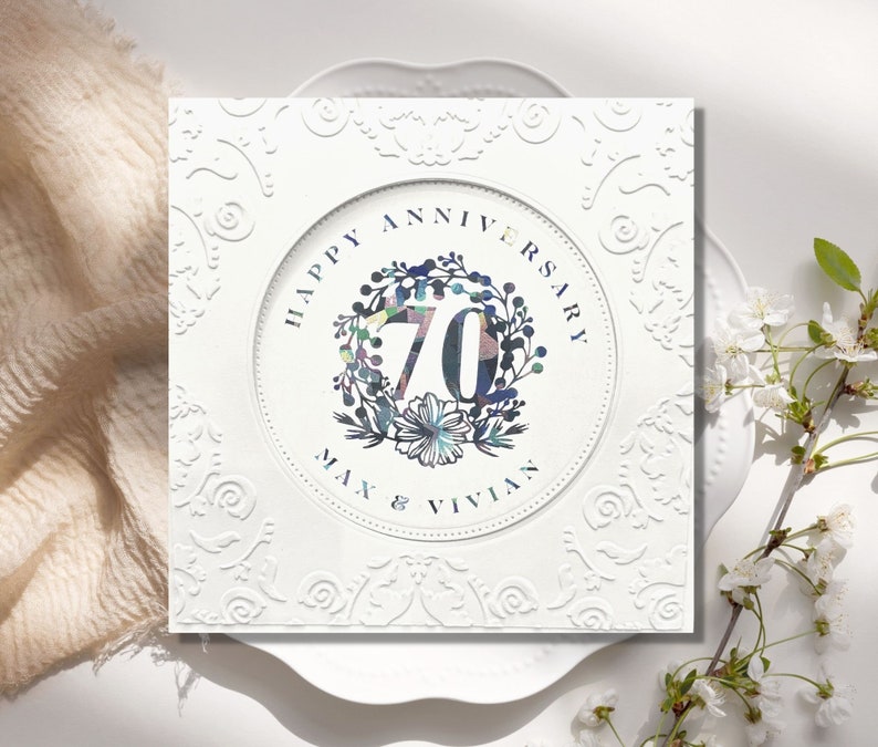 May include: White card with a decorative border and a circle in the center. The circle has a floral wreath with the number 70 in the center. The text "Happy Anniversary Max & Vivian" is below the wreath.