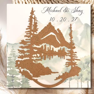 May include: A square wedding invitation with a mountain and tree design. The invitation features a brown cutout of a mountain and trees over a watercolor background. The text "Michael & Shay" and the date "10.20.27" are written in script.