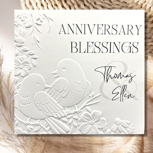 May include: White card with embossed floral design and two birds. The text "Anniversary Blessings Thomas & Ellen" is printed in black.