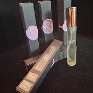 May include: A clear glass perfume bottle with a gold cap and the text "AMETHYST BLOOM". The bottle is next to several dark gray boxes with a purple wax seal featuring a rabbit design. The boxes have the text "15ml" and "SHAKE WELL".