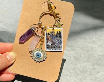 May include: Gold keychain with a tarot card charm featuring The Empress, a pink and purple crystal charm, and a blue evil eye charm.