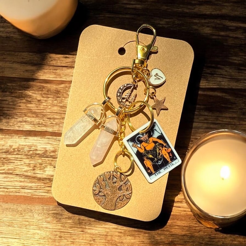 May include: A gold keychain with various charms, including two crystal pendants, a tree of life charm, a star, a heart, and a tarot card that reads "THE DEVIL". The keychain is displayed on a brown card.