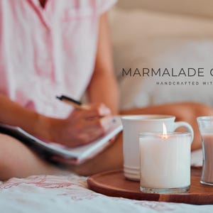 May include: A cozy scene with lit candles and a mug on a wooden tray. The image features the text "MARMALADE OFFICIAL HANDCRAFTED WITH LOVE". The candles are in glass containers, and the scene is set on a bed.