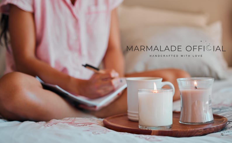 May include: A cozy scene with lit candles and a mug on a wooden tray. The image features the text "MARMALADE OFFICIAL HANDCRAFTED WITH LOVE". The candles are in glass containers, and the scene is set on a bed.