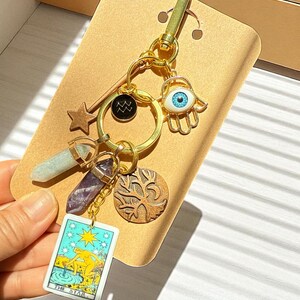 May include: Gold-toned keychain with various charms, including a star, a crystal, a tarot card, and a tree of life. The tarot card displays "THE STAR" with a celestial illustration. Other charms include an eye and a zodiac symbol.
