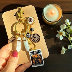 May include: A gold keychain with various charms, including a tarot card, crystals, and an eye pendant. The tarot card depicts a devil figure. The keychain is displayed on a brown card with a lit candle and flowers in the background.