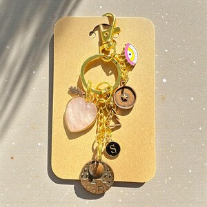 May include: A gold-coloured keyring with various charms. Charms include a pink heart-shaped stone, a compass, a small star, a black circle with the letter "S", and an eye-shaped charm. The keyring is attached to a tan-coloured card.