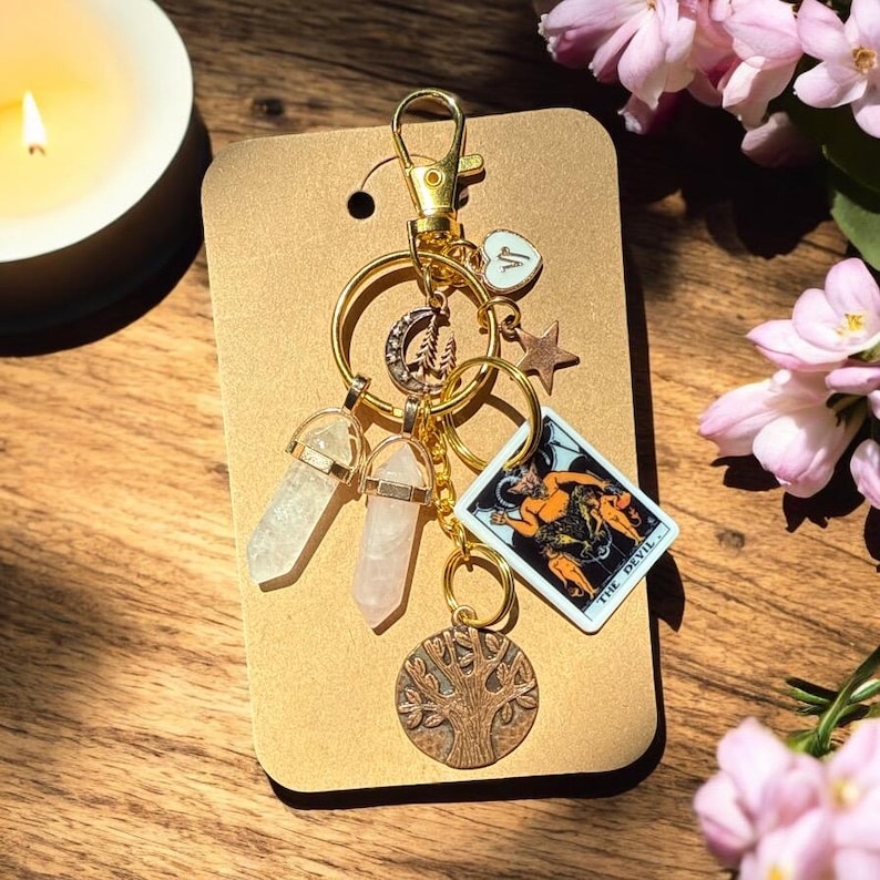 May include: Gold-toned keychain with multiple charms, including two crystal pendants, a crescent moon, a star, a tree of life, and a tarot card. The tarot card features an illustration of The Devil. The keychain is attached to a brown card.