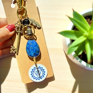 May include: A gold-toned keychain with various charms, including a key, a feather, a bell, and a blue stone. The keychain also features a white and blue circular charm with a geometric design. The background is a light brown card.